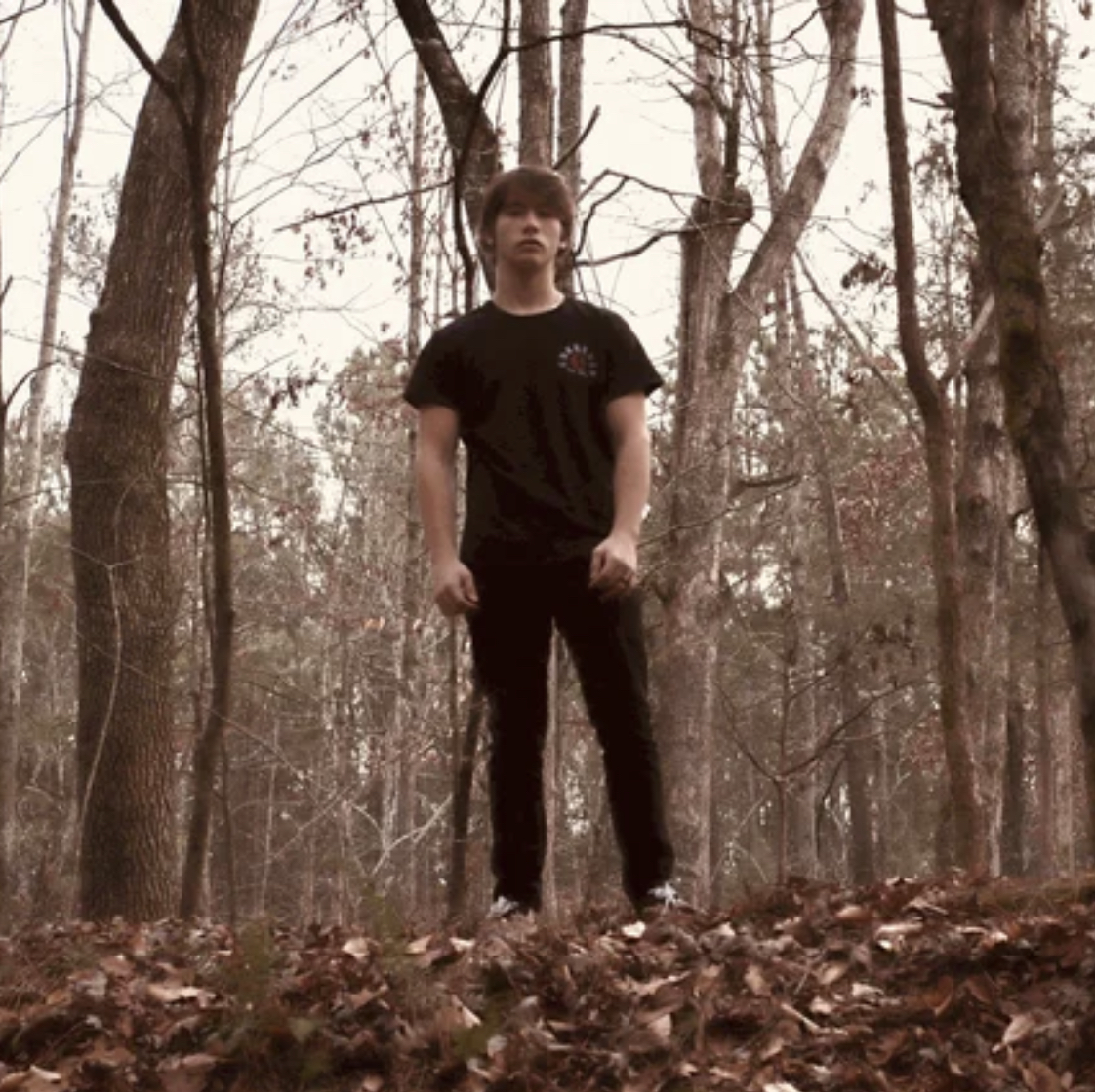 Young man standing in a forest with trees and fallen leaves, wearing a black t-shirt and pants.