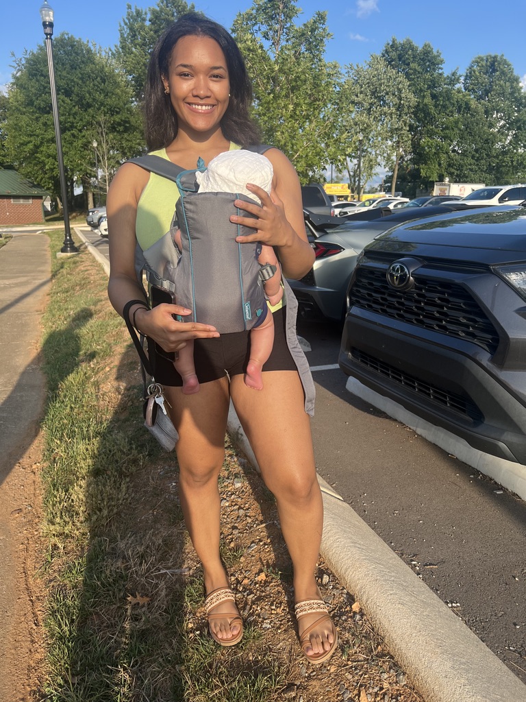 Smiling woman holding a baby in a gray carrier outdoors on a sunny day, with parked cars and trees in background.