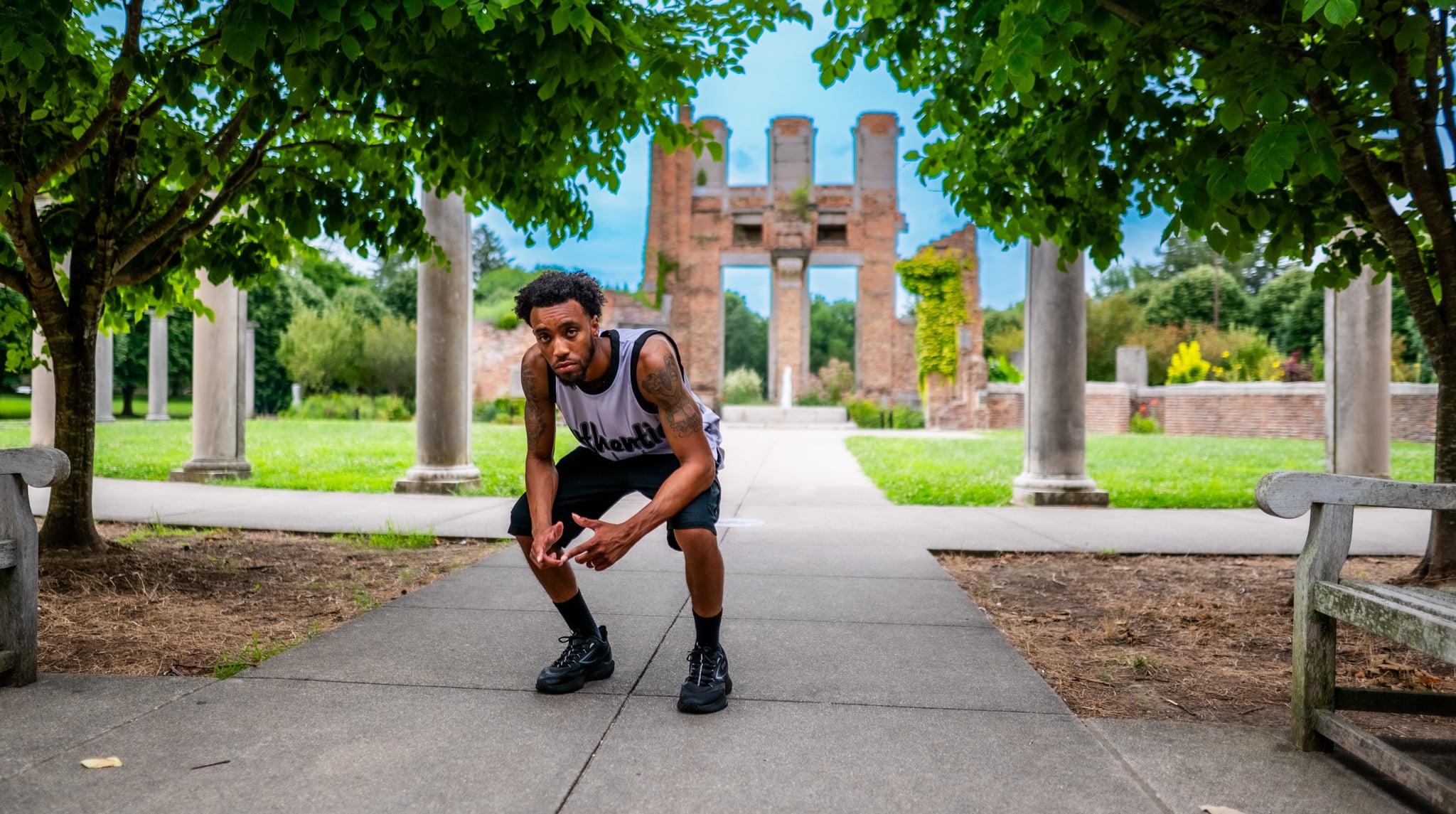Young man crouching on a pathway in a park with columns and trees in the background.