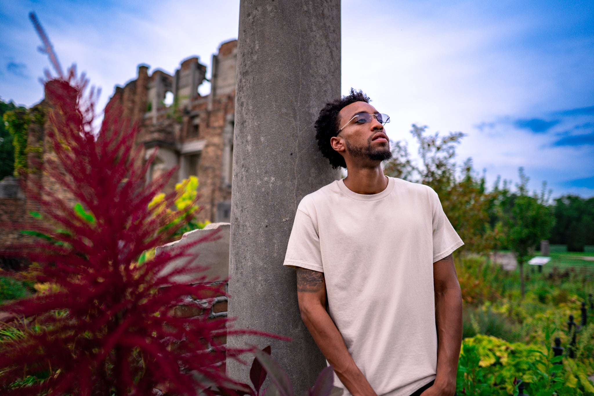 Young man with curly hair and glasses leaning against a concrete pillar outdoors, with colorful plants and ruins in background.