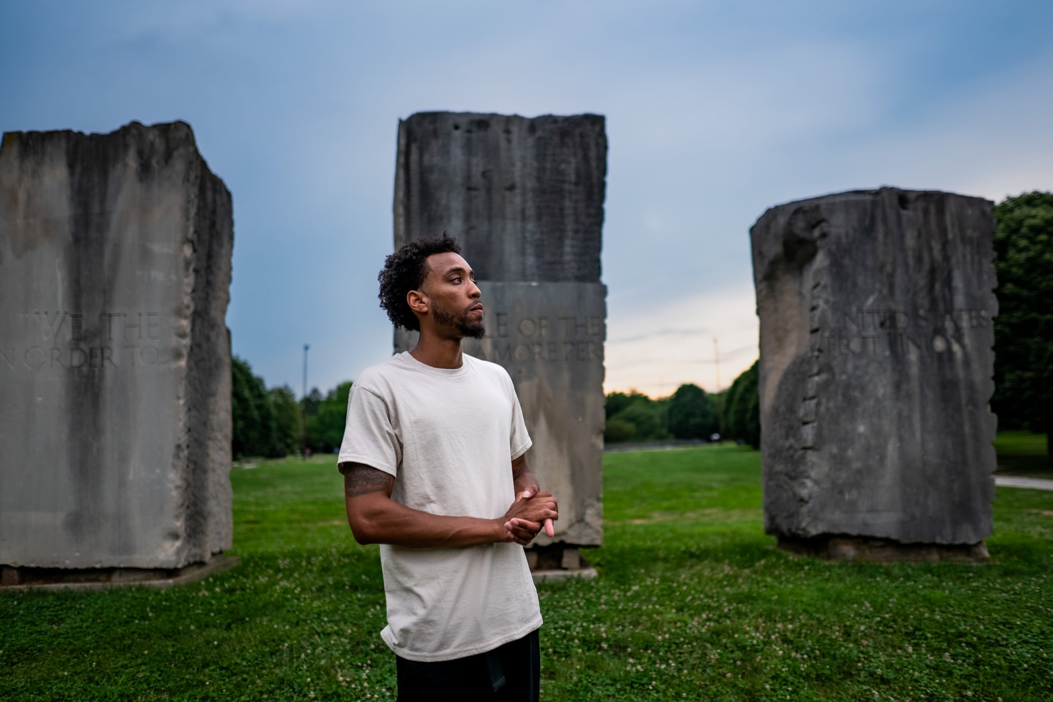 Young man with curly hair and beard stands outdoors among large concrete blocks on grass, looking to the side.