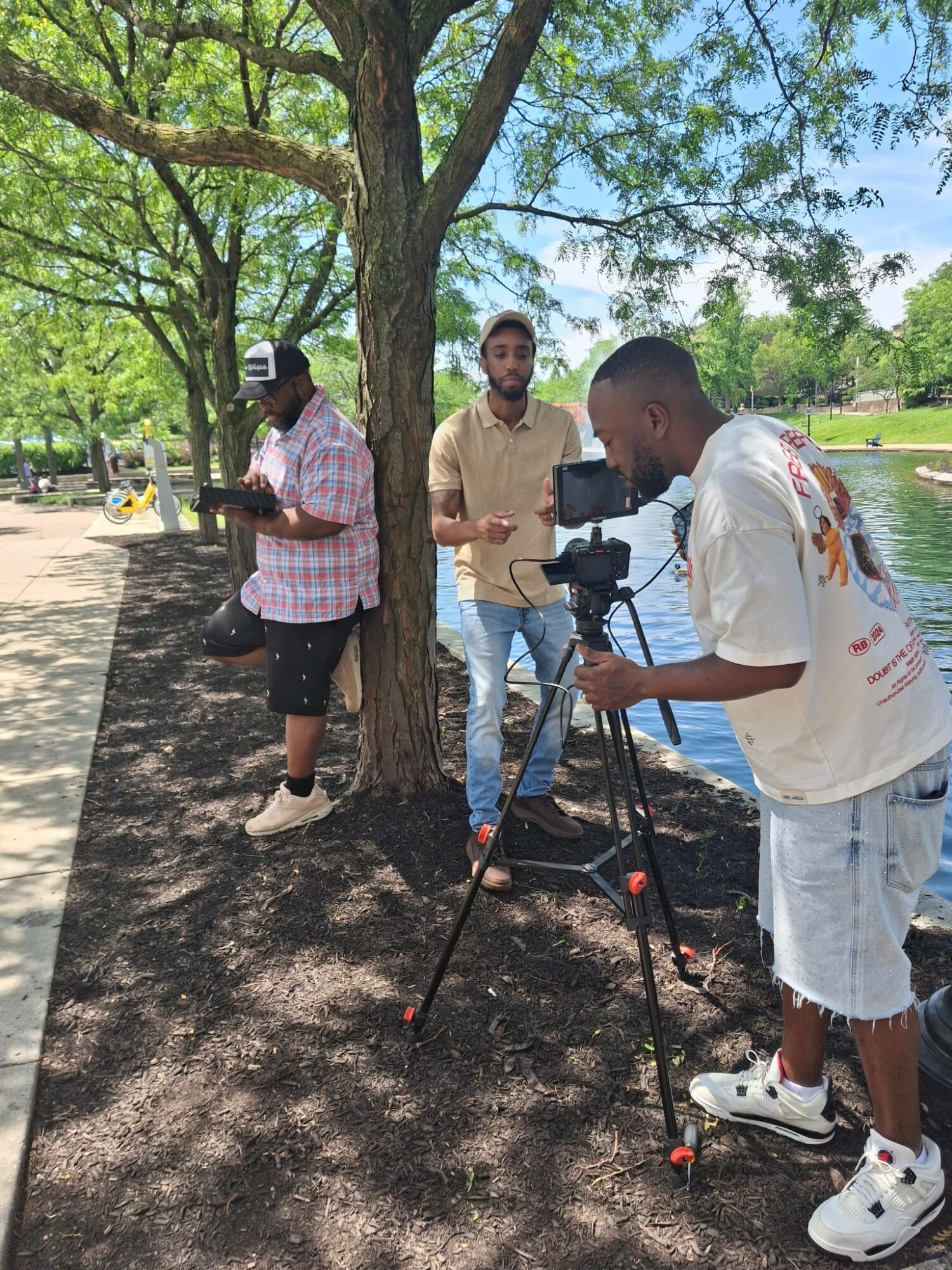Three people near a tree by a lake, one adjusting a camera on a tripod, others standing nearby, sunny day.