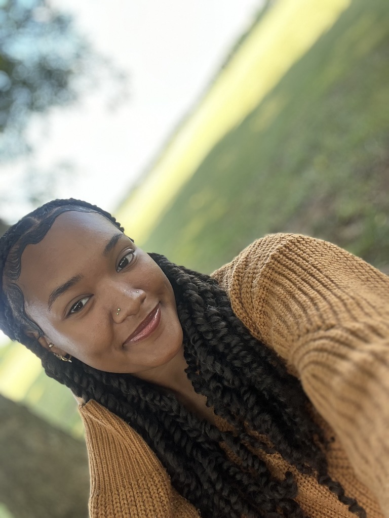 Young girl with braided hair smiling outdoors, wearing a brown sweater, in a grassy park with trees in the background.