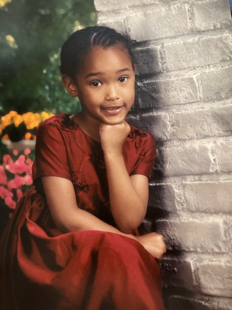 Young girl in a red dress sitting against a brick wall, smiling with her chin resting on her hand.