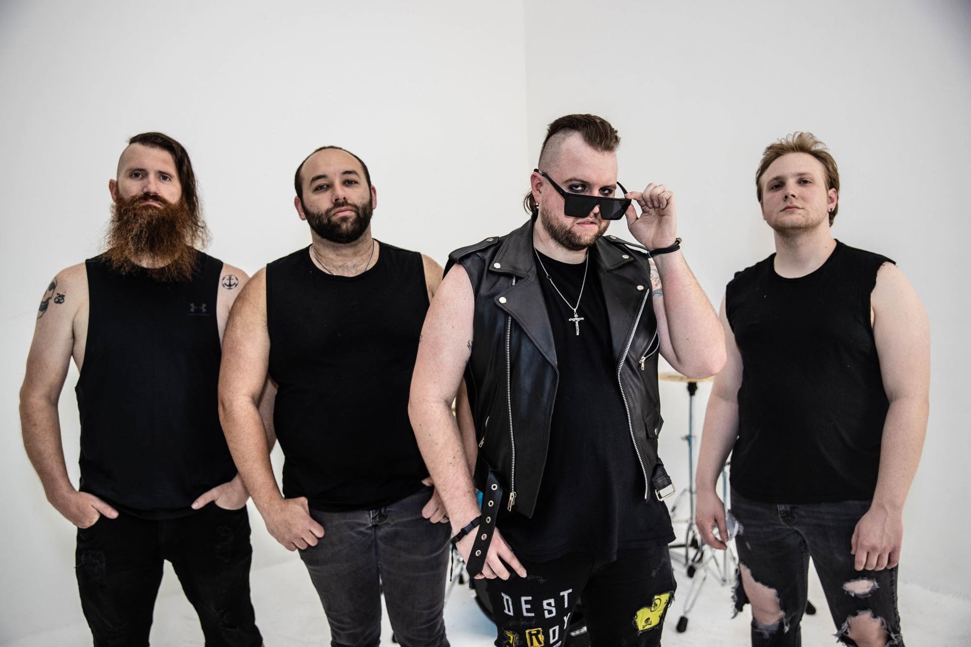Four men standing against a plain background, wearing black sleeveless shirts, with one man in sunglasses and a leather vest.