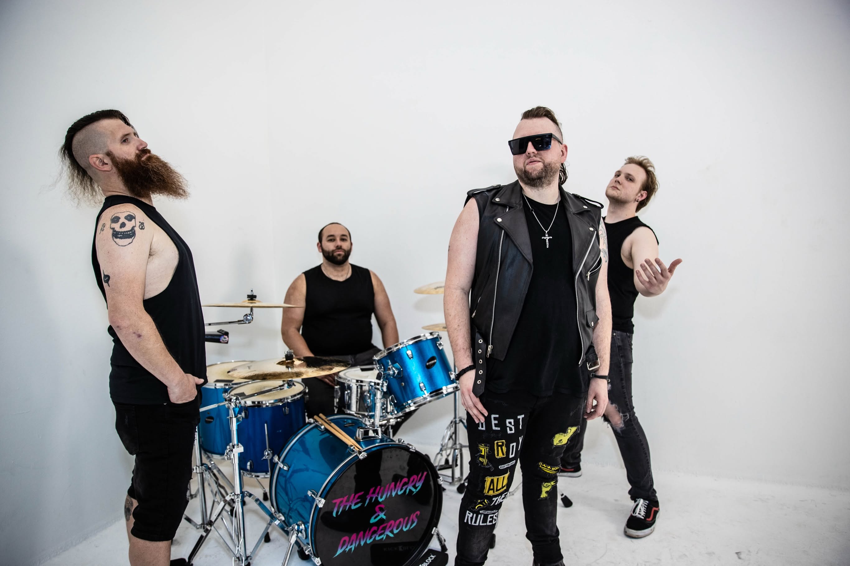 Four band members with musical instruments in a white studio, dressed in black, some wearing sunglasses, posing confidently.