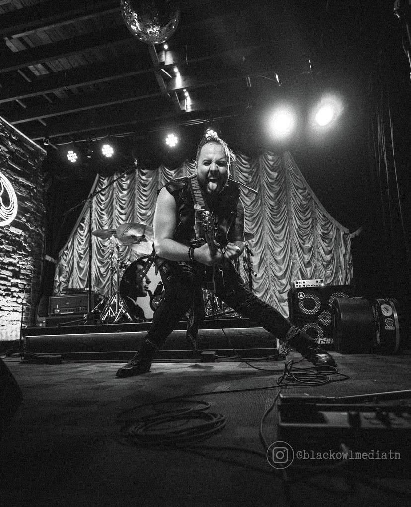 Performer singing on stage with microphone, stage lights shining, curtain backdrop, and musical equipment visible.