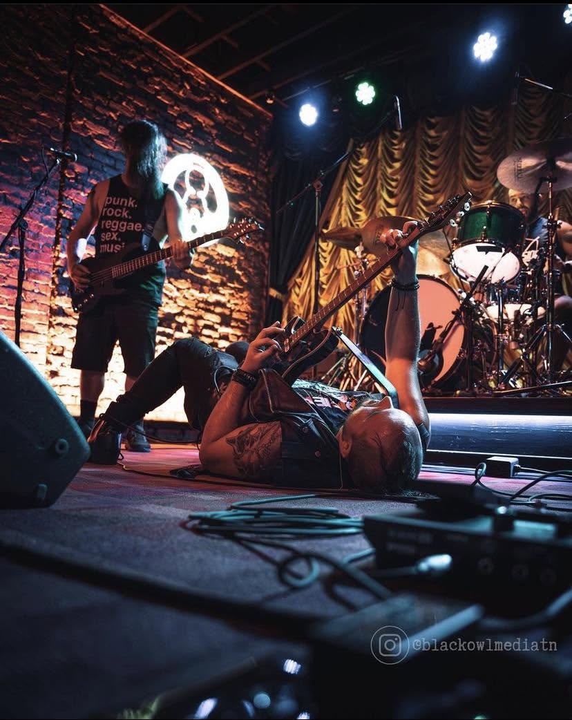Person lying on stage with legs raised, holding a microphone, with band members and stage lights in background.