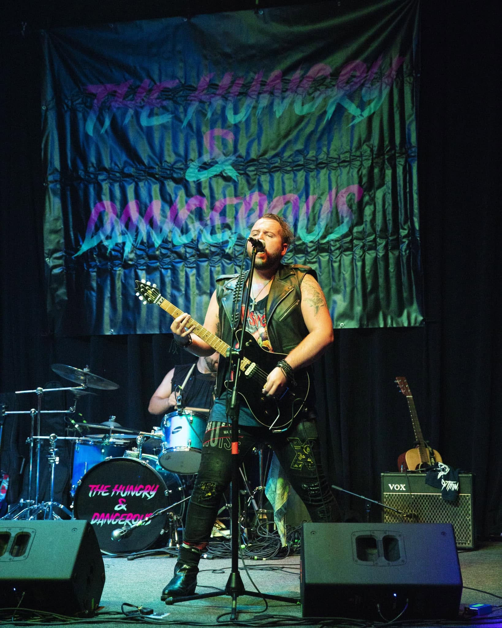 Musician with a guitar performing on stage with a drum set and banner in the background.