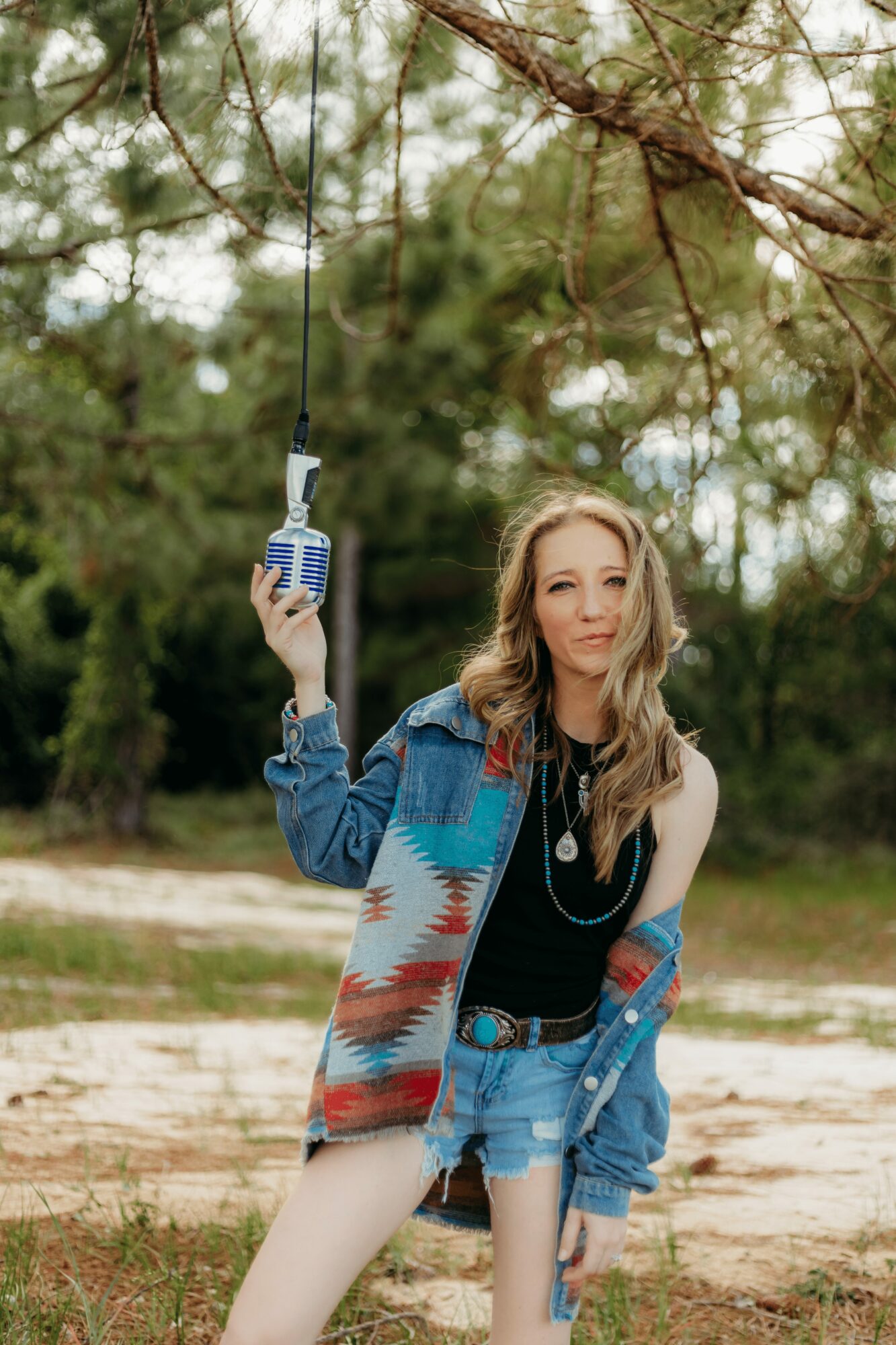 Woman standing outdoors holding a radio, wearing a colorful jacket and denim shorts, with trees in the background.