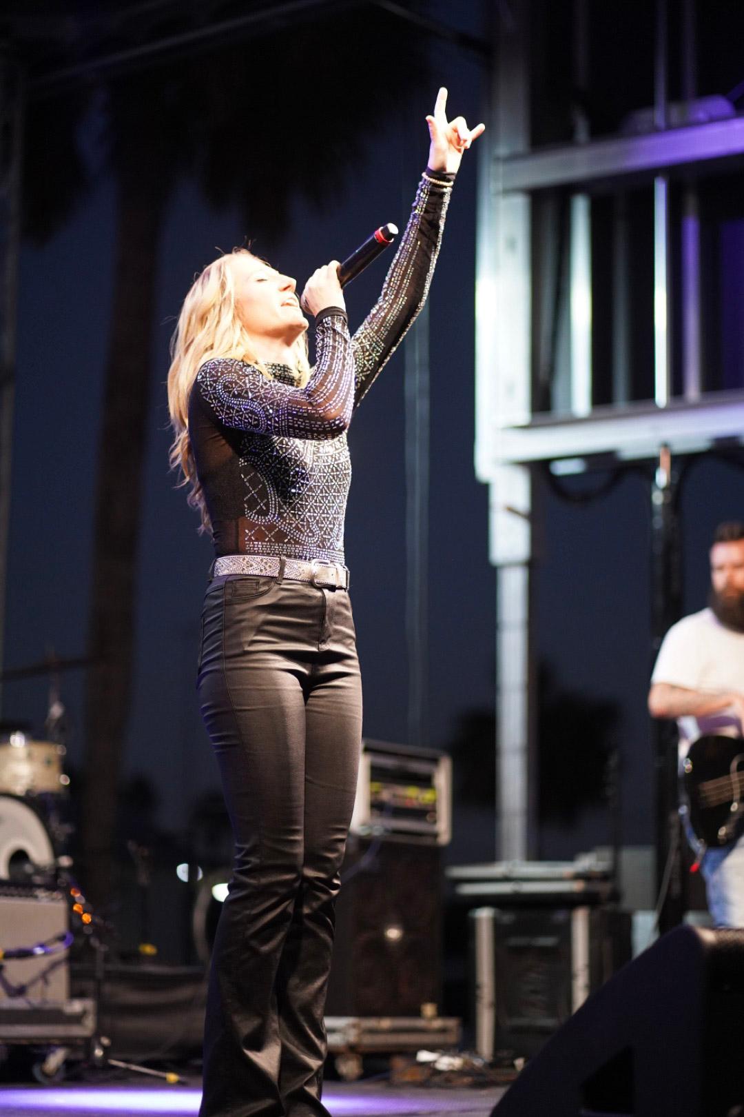 Female singer with blonde hair performs on stage, holding a microphone and raising her hand, with band members in background.