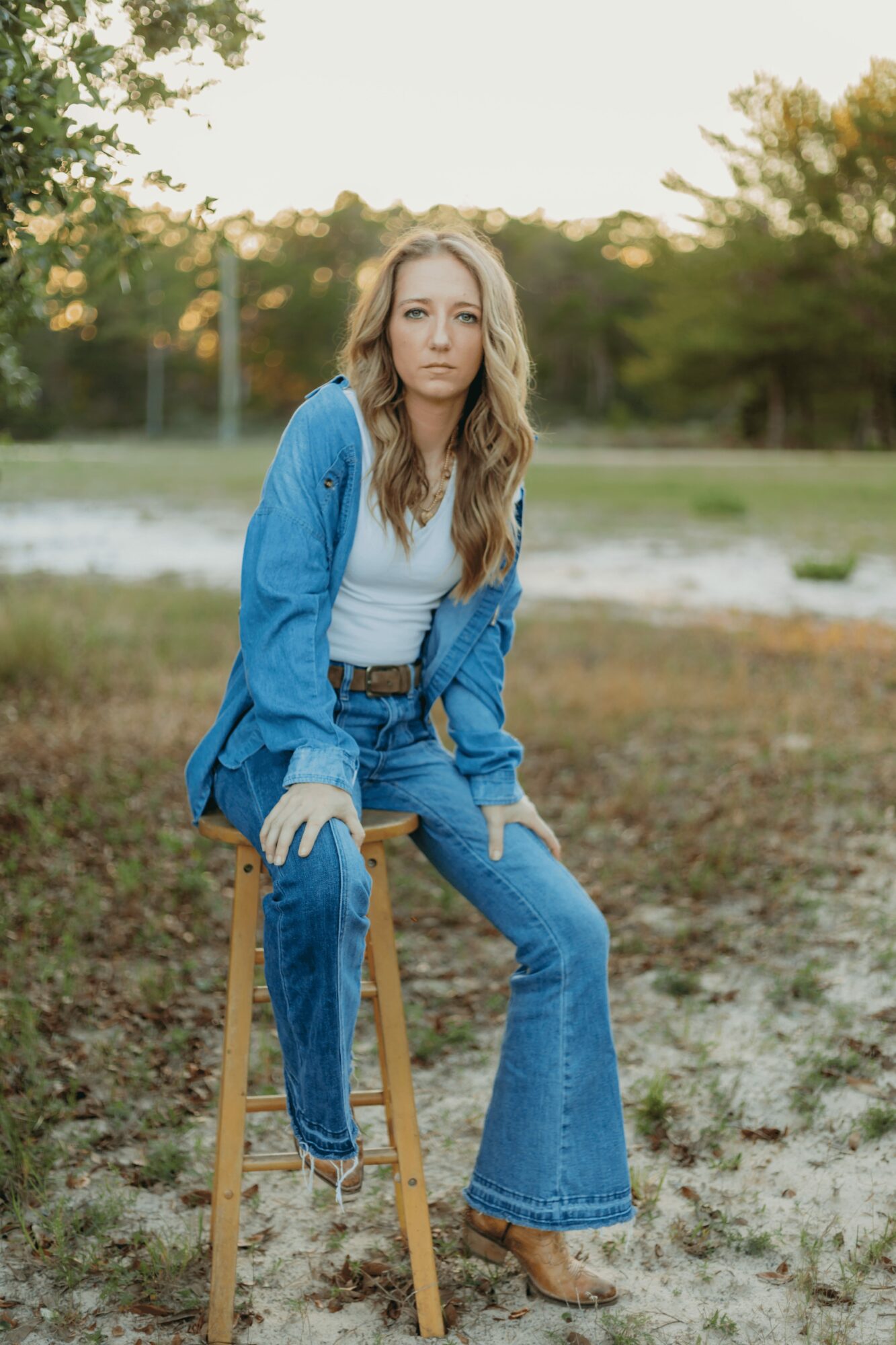 Young woman with long hair sitting on a wooden stool outdoors, wearing a blue jacket and jeans, with trees in the background.
