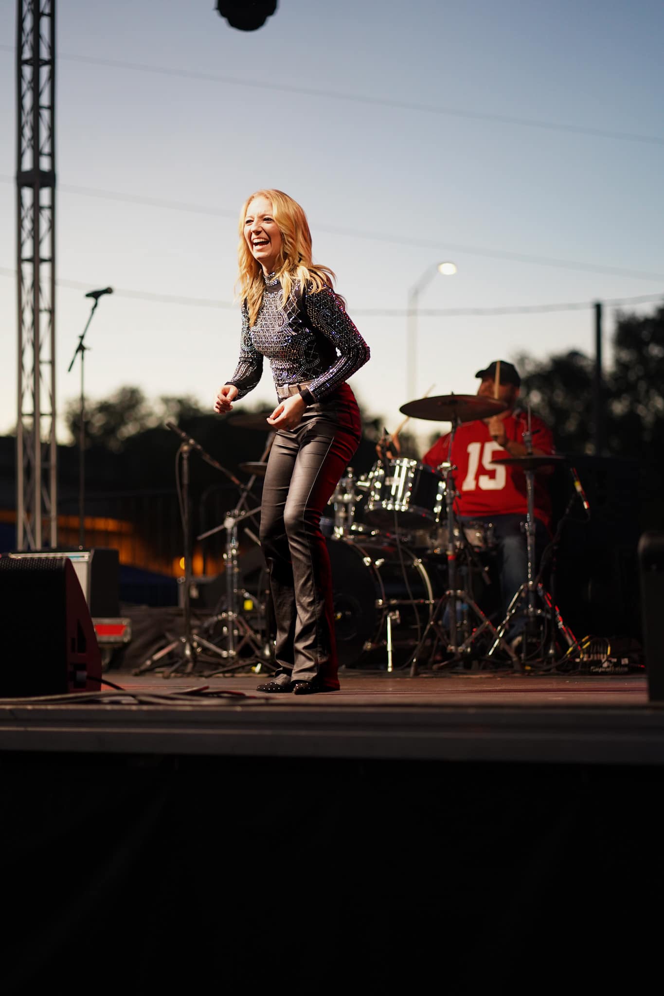 A woman with blonde hair singing on stage during an outdoor concert, with a drummer in the background wearing a red shirt.
