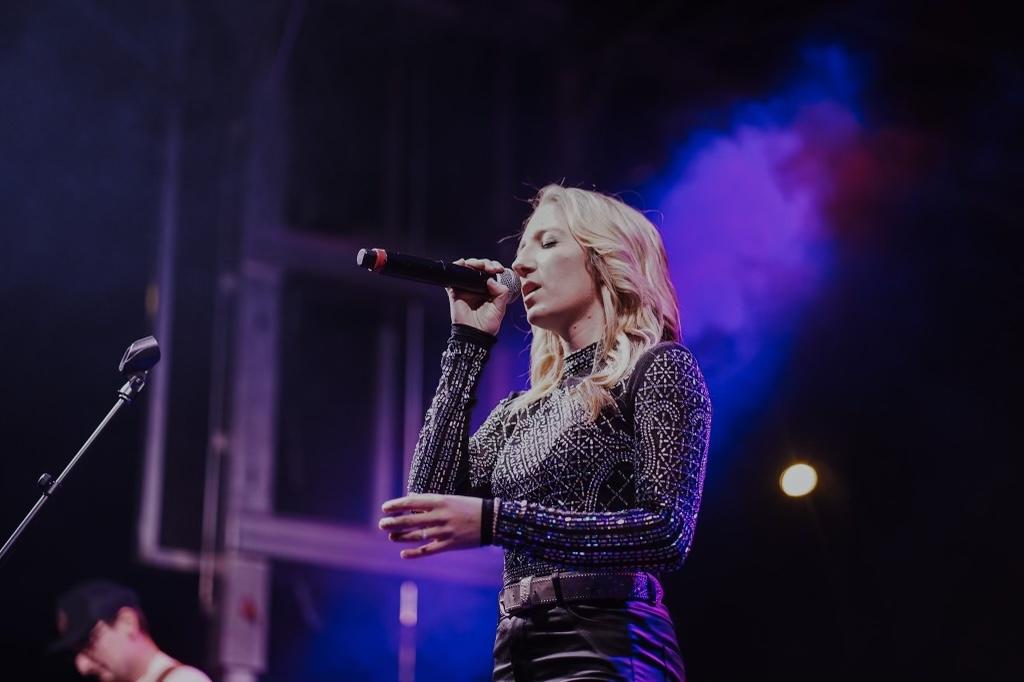 A woman with blonde hair singing into a microphone on stage with colorful lighting.