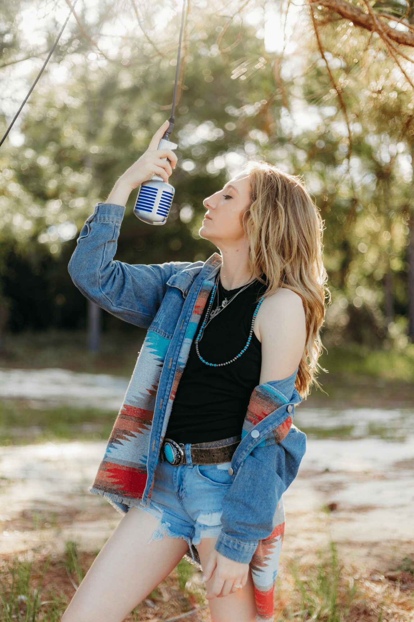 Young woman with long hair, wearing a black top and denim shorts, holding a soda can to her mouth outdoors.