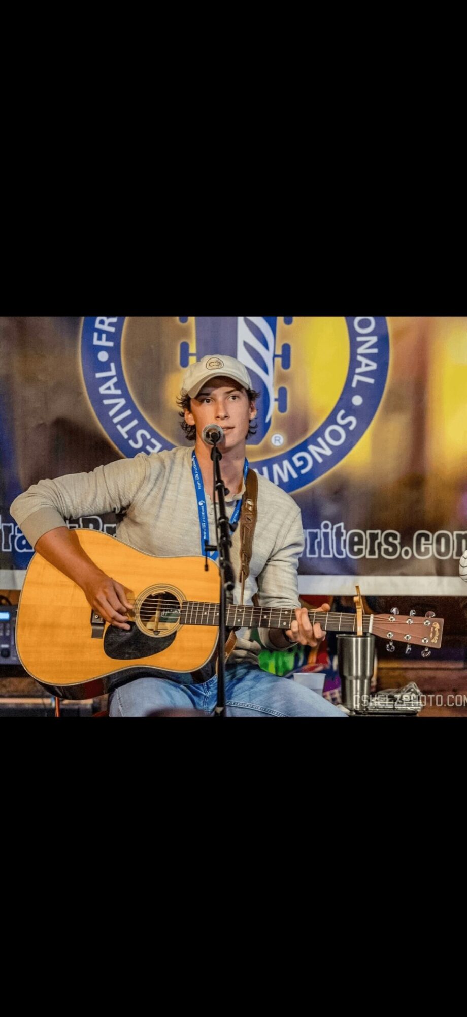 Person playing guitar and singing into a microphone, wearing a hat, with a blue circular background and a banner.