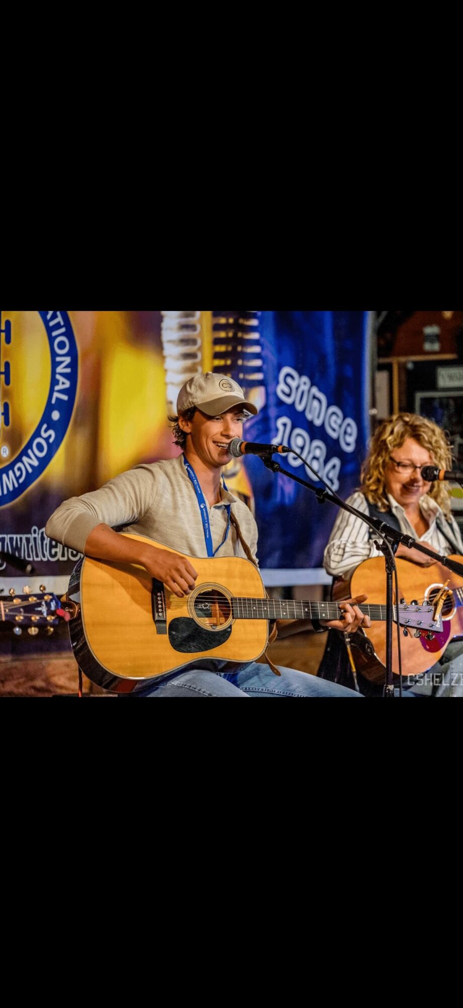 Two musicians playing guitars and singing into microphones on stage, with a blue banner in the background.