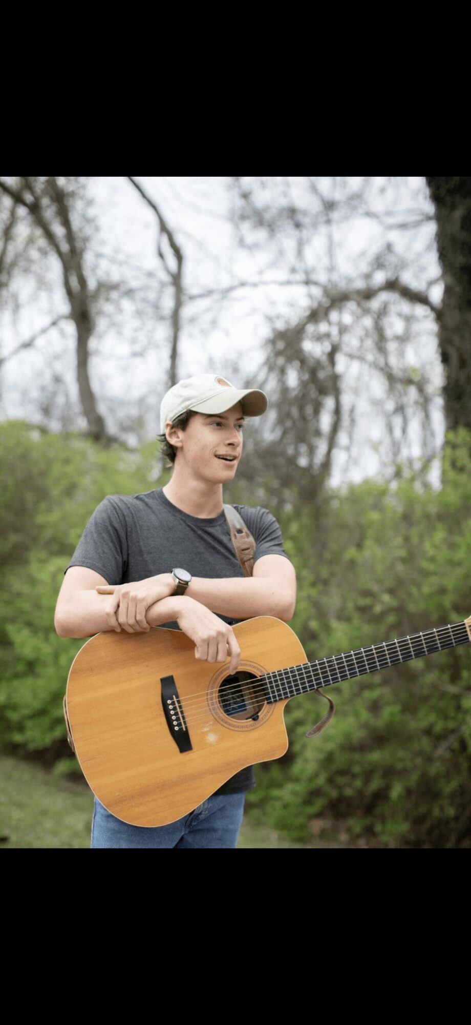 Person holding an acoustic guitar outdoors, wearing a cap and casual clothing, with trees in the background.