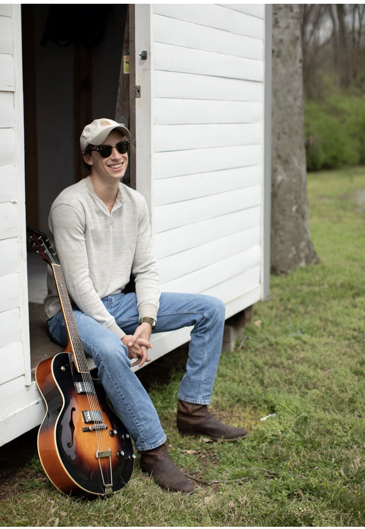 Person sitting outside a white shed with a guitar, wearing sunglasses, a hat, a light sweater, and jeans.