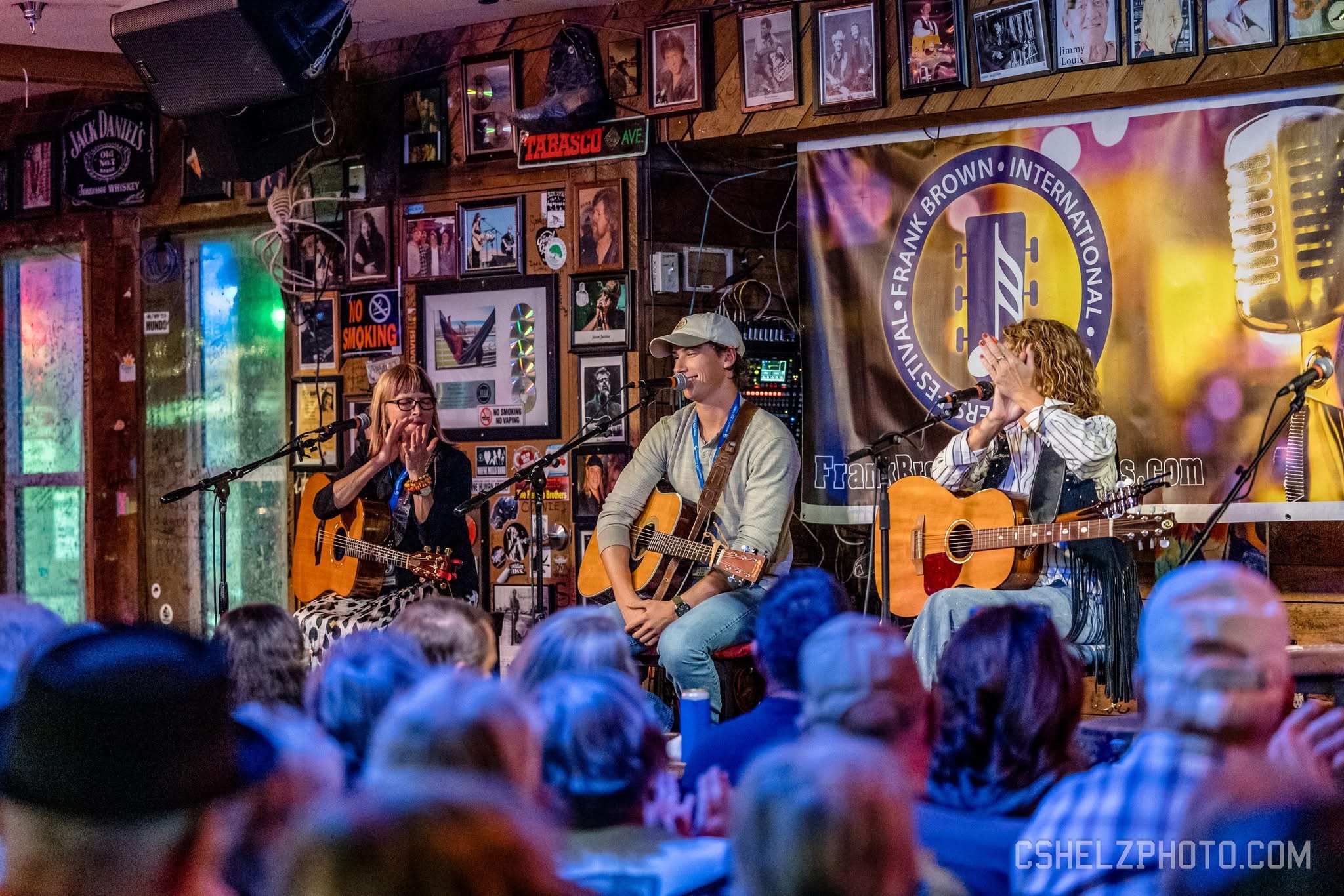 Three musicians perform on stage with guitars, a woman singing, and a man wearing a hat, in front of an audience.