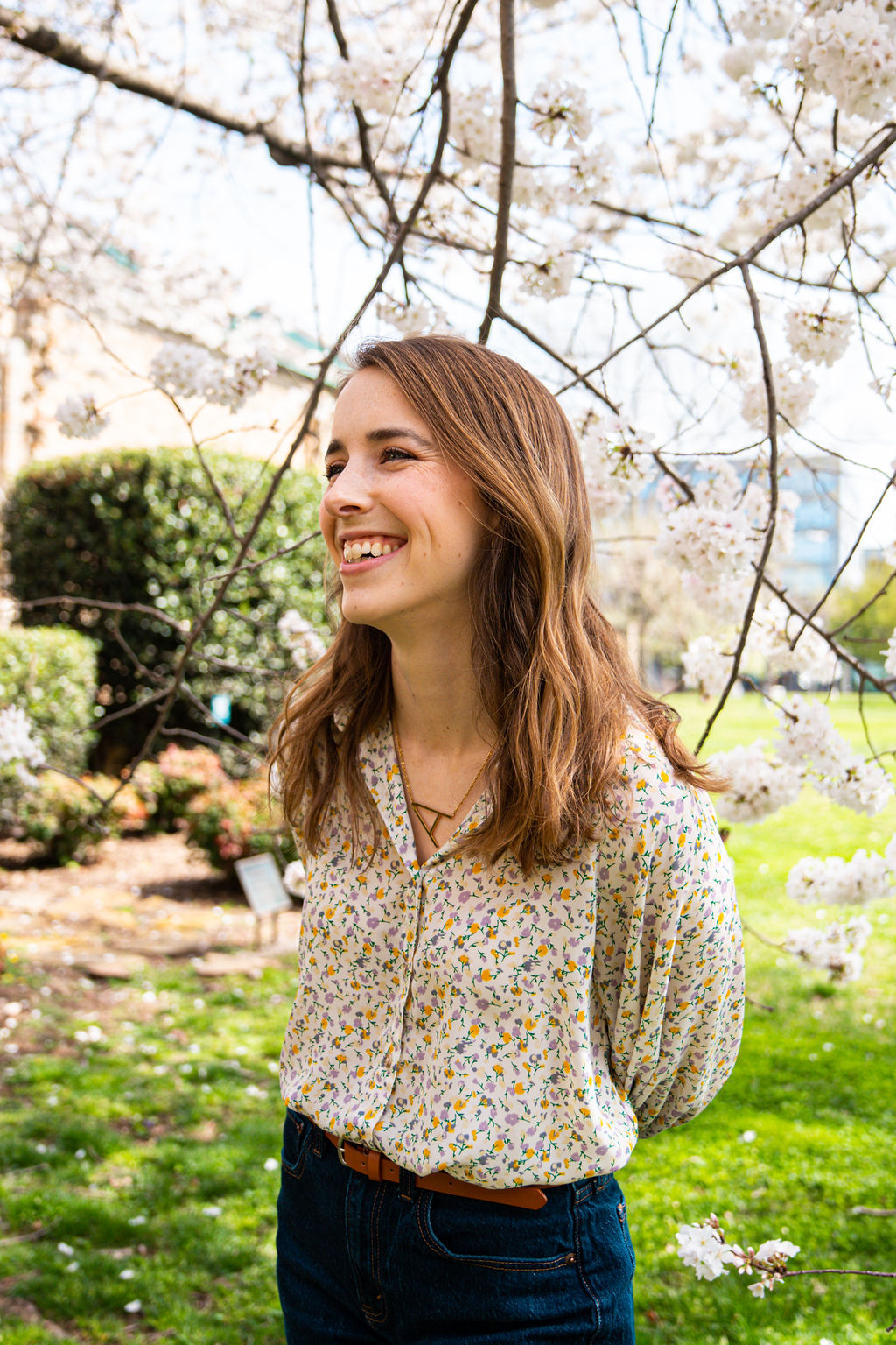 Young woman smiling outdoors under blooming cherry blossom tree, wearing a floral blouse and jeans.