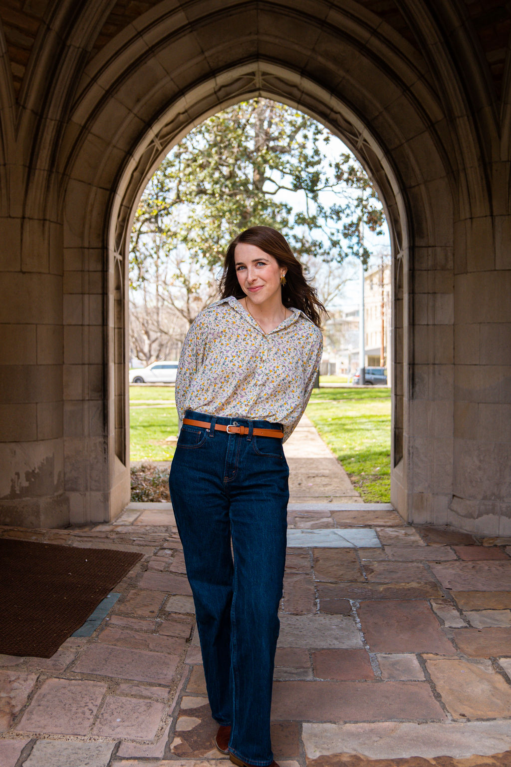 Young woman with long dark hair standing in an arched stone doorway outdoors, smiling, with trees and grass behind her.