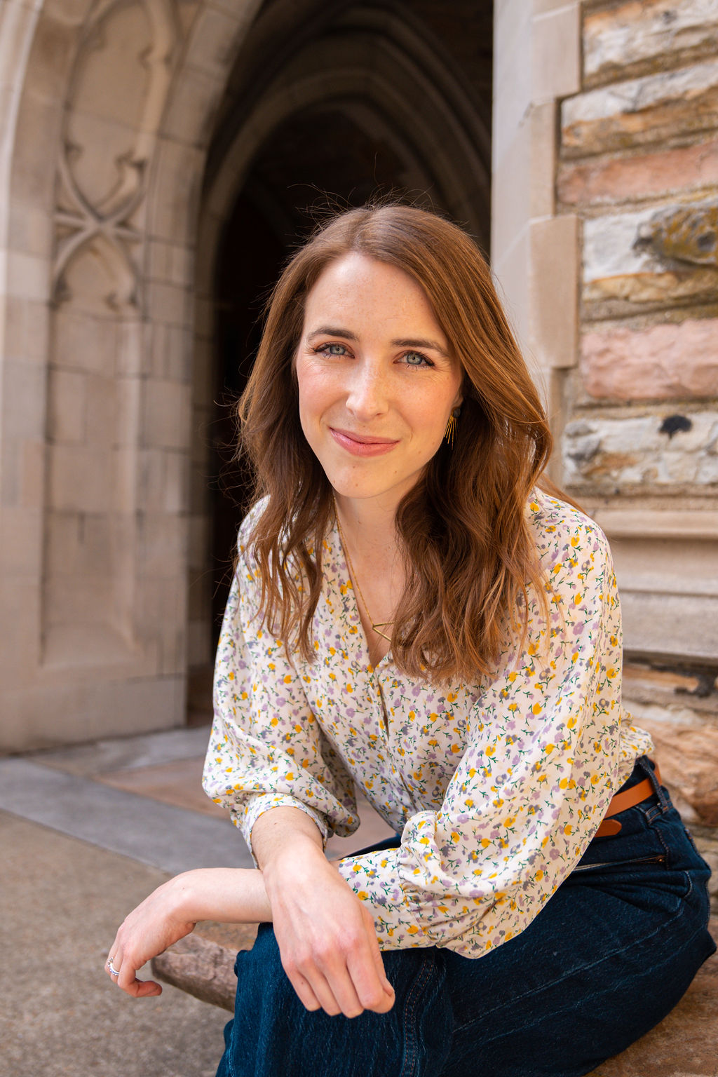 Young woman with long brown hair smiling, sitting outdoors near a stone building and archway, wearing a floral blouse and dark jeans.