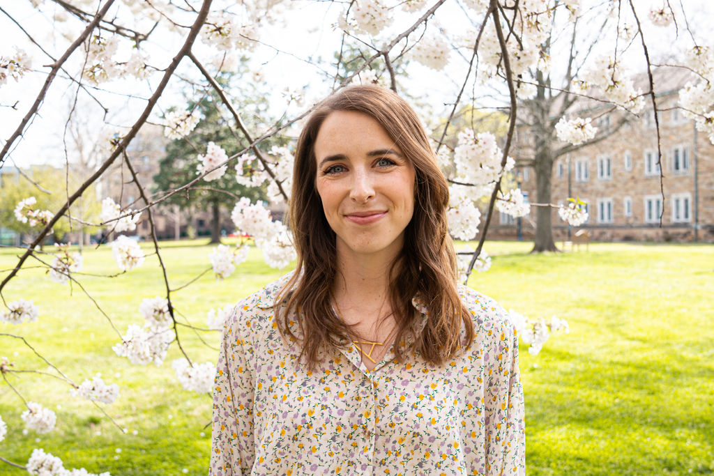 Woman with long brown hair smiling outdoors near blooming trees and a grassy area.