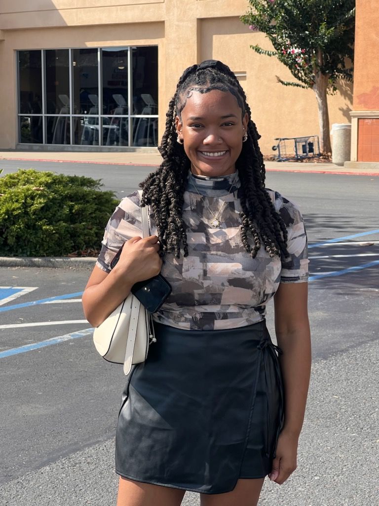 Young woman with braided hair smiling, wearing a camouflage top and black skirt, standing outdoors near a building and greenery.