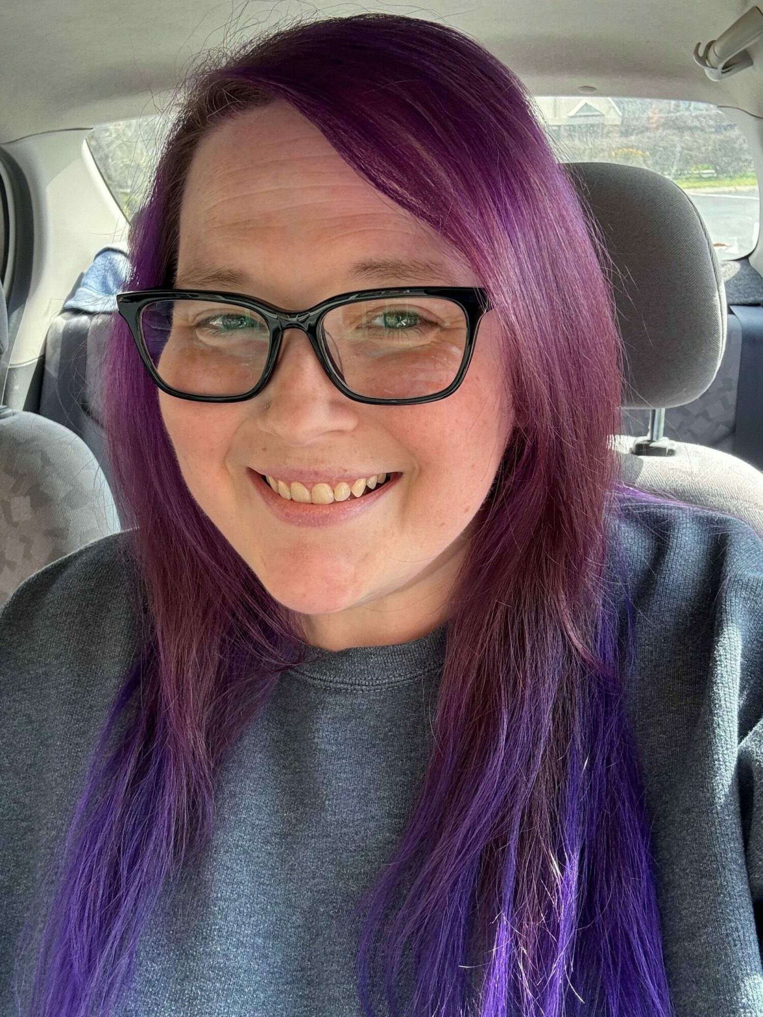 Woman with purple hair and glasses smiling inside a vehicle, background shows car interior and window.