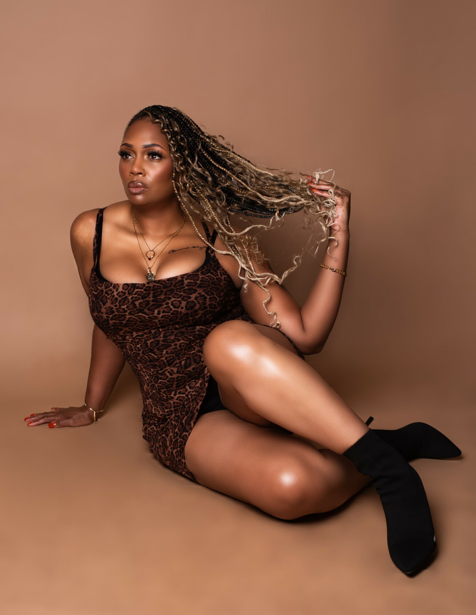 Woman with long hair sitting on the floor, wearing a dress and black boots, against a brown background.