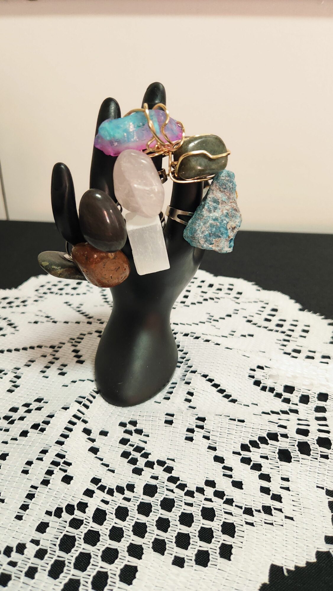 Black hand-shaped jewelry holder with various colorful rings and stones, placed on a white lace surface.