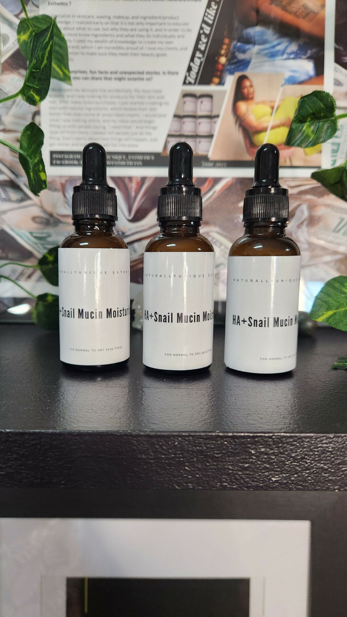 Three white bottles with black caps on a black surface, with plants and magazines in the background.