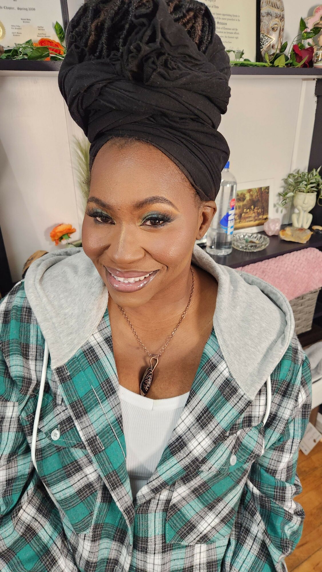 Woman with a headwrap smiling, wearing a plaid shirt and a necklace, in a room with flowers and pictures.