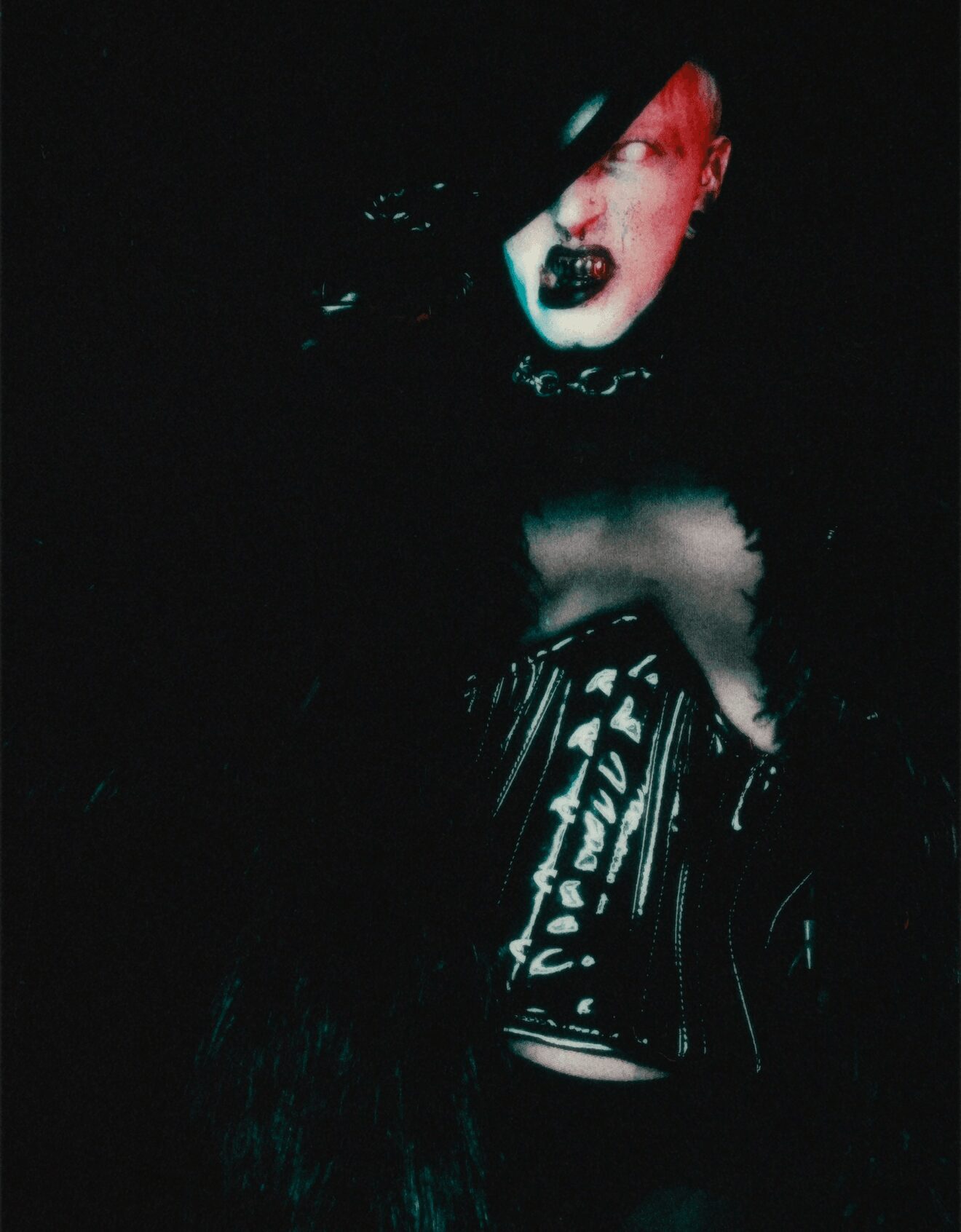 Person with red hair and dark makeup, wearing a black outfit, in a dark setting.