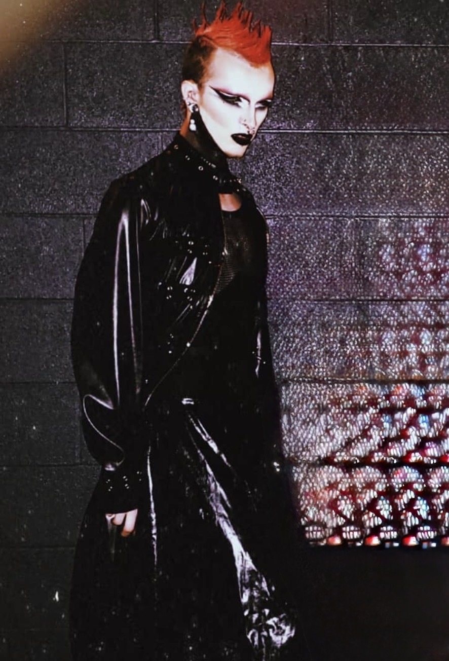 Person with bright red hair, dark makeup, wearing a shiny black outfit, standing against a textured wall.