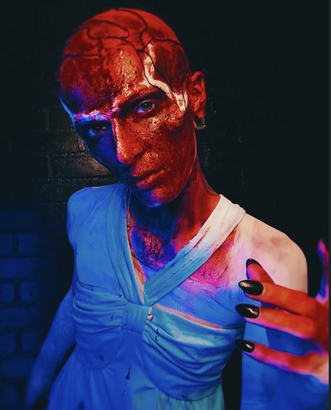 Person with face and neck covered in red paint, wearing a blue top, taking a selfie with painted hand, dark background.