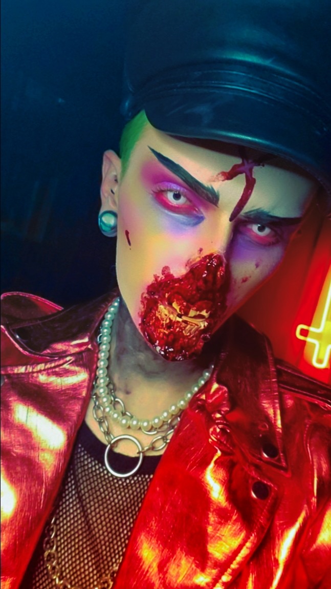 Person with colorful makeup, black hat, red shiny jacket, and pearl necklace, with a dark background and neon light.