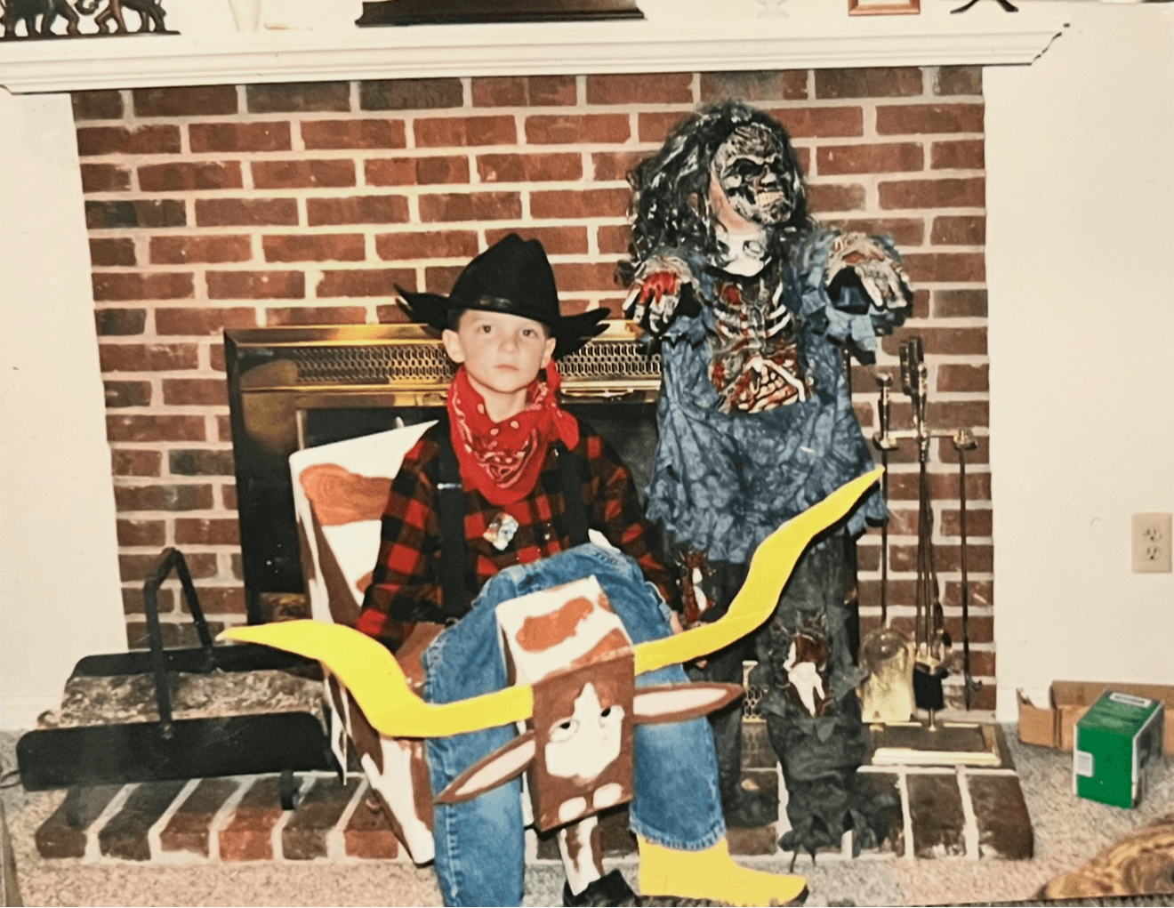 Child dressed as a cowboy with a red bandana, sitting in a cardboard horse costume, in front of a brick fireplace with a witch decoration nearby.