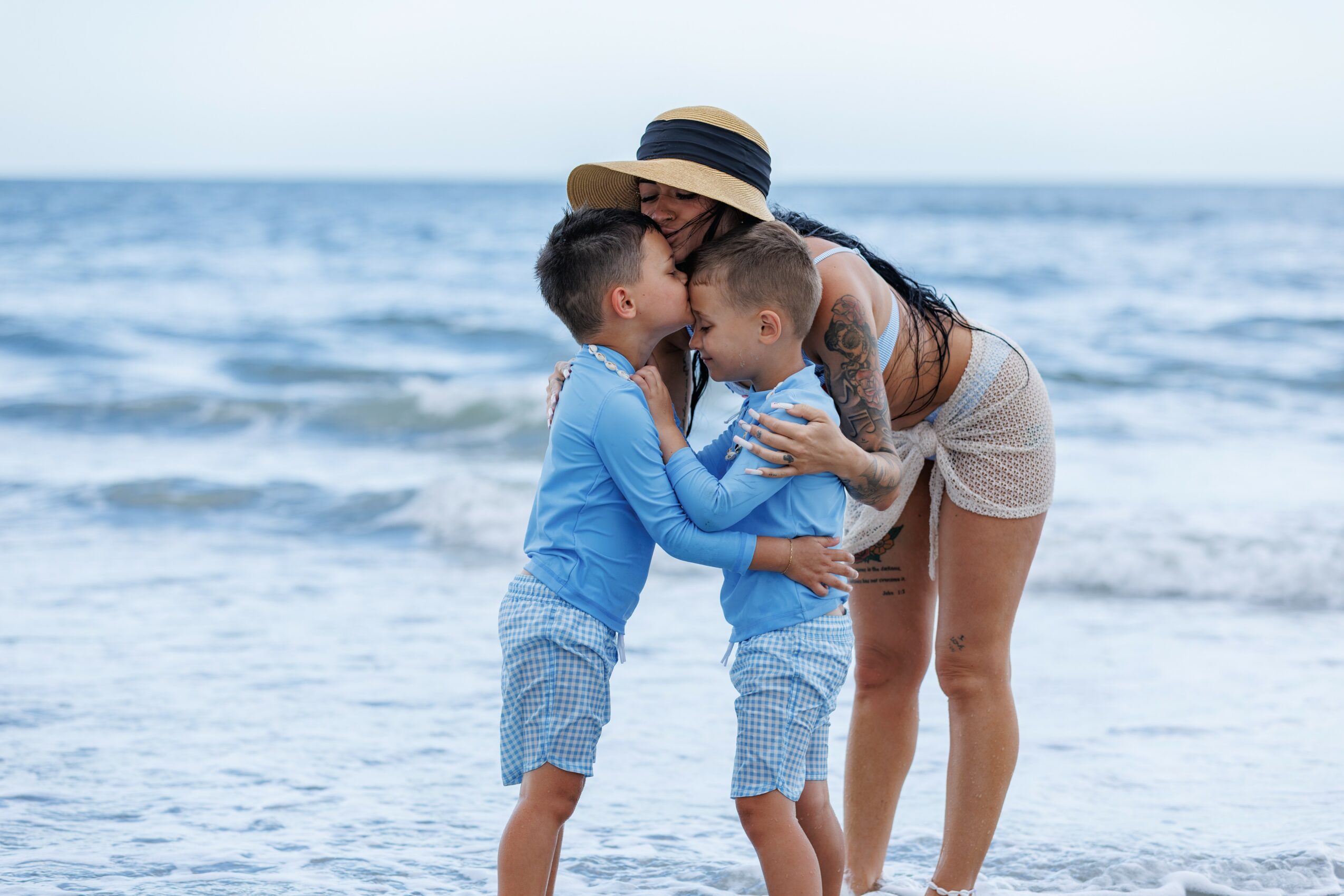 Woman with two children on the beach, kissing each other, ocean in background, woman wearing hat and shorts.