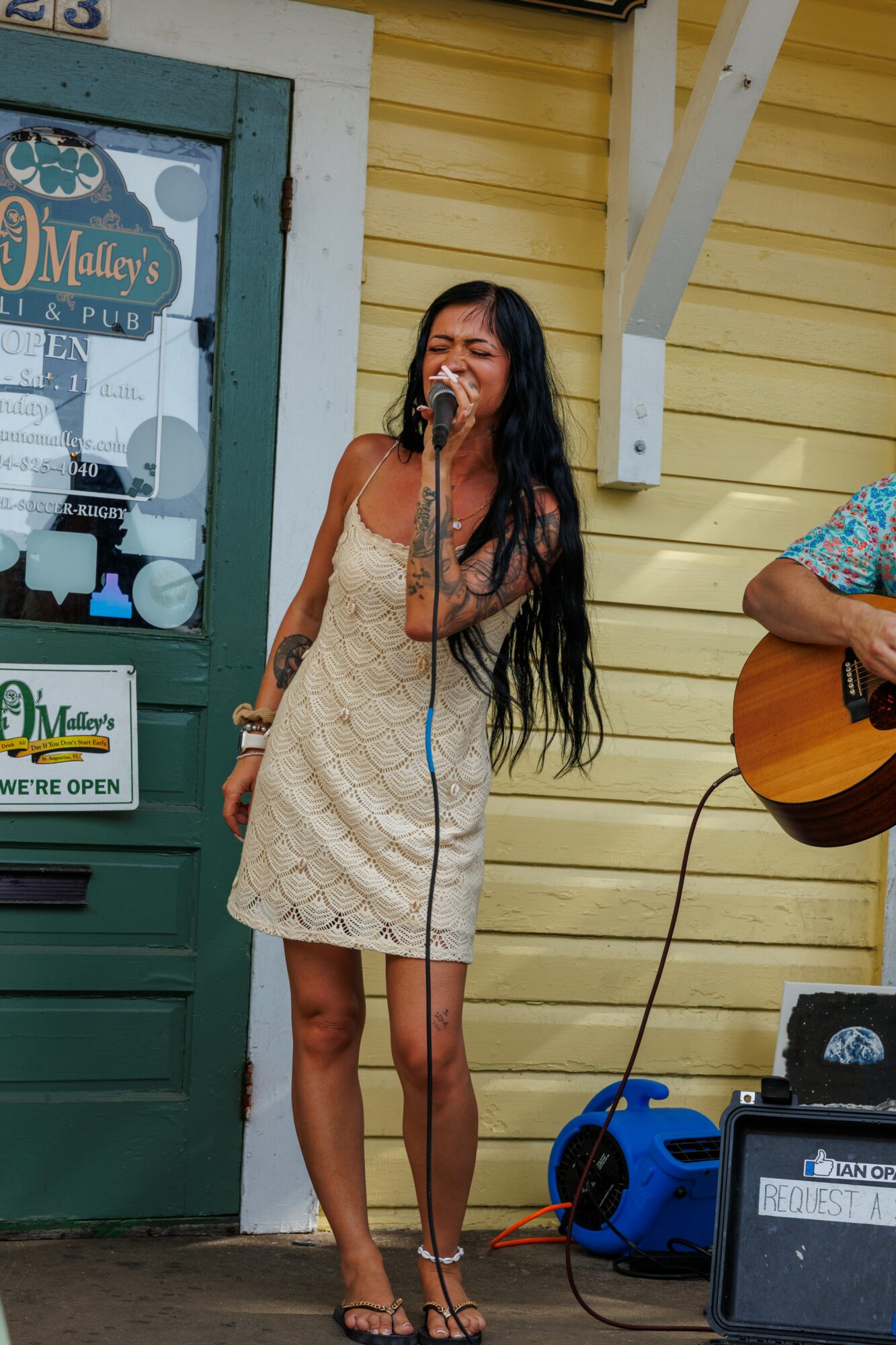 Woman singing into a microphone outdoors, with a person playing guitar beside her, in front of a yellow building.