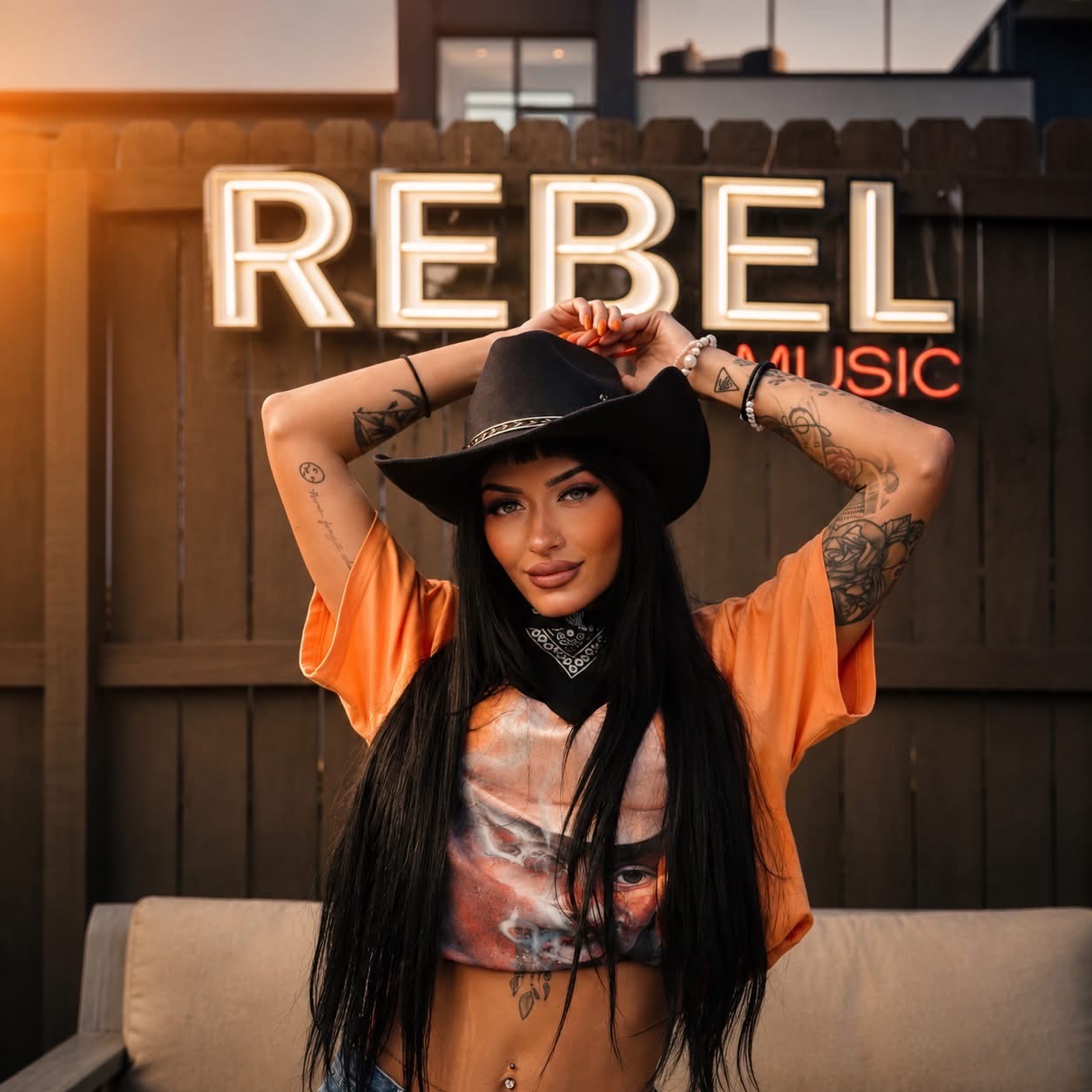 Woman with long dark hair wearing a black hat and orange shirt, standing in front of a wooden fence with a neon sign that says REBEL and MUSIC.