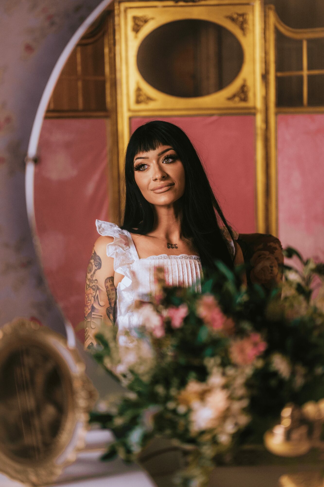 Woman with long black hair and lace dress looking at mirror, pink and gold background, floral arrangement in foreground.