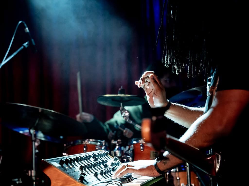 Person playing drums on stage with dark background, stage lighting, and musical equipment visible.