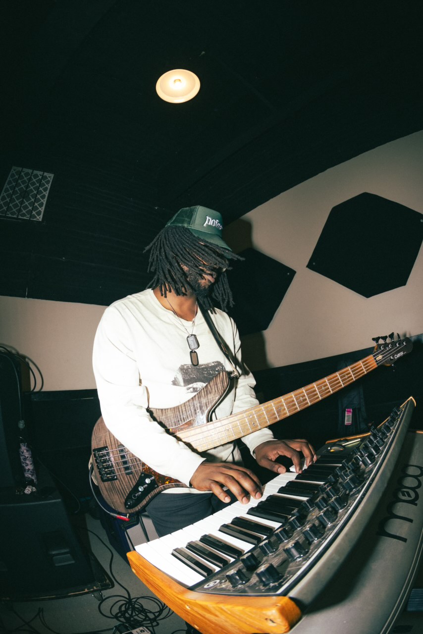 Person with dreadlocks wearing a cap and white shirt playing a guitar and keyboard in a dark room.