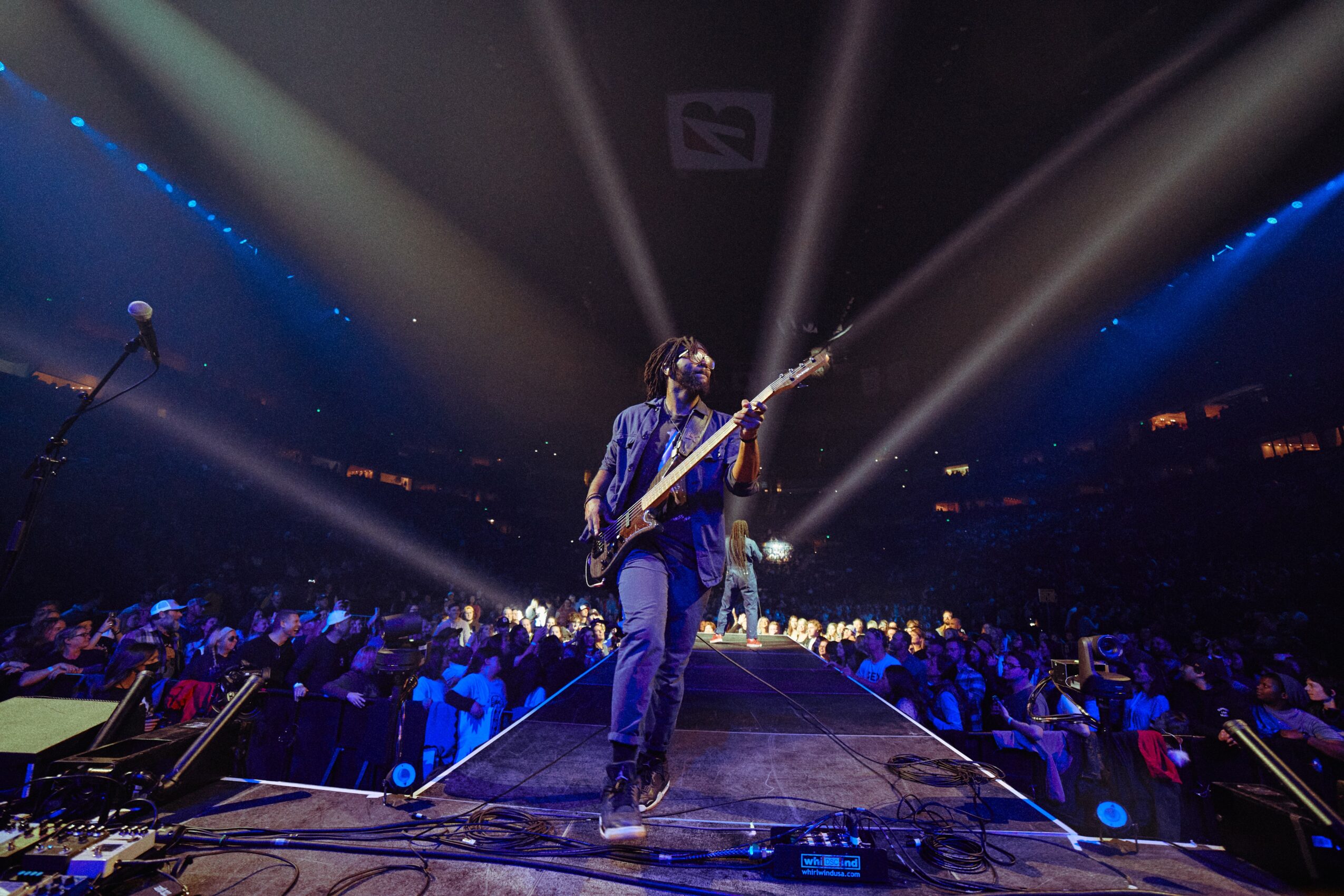 Musician playing guitar on stage with bright spotlights, audience in background, concert setting, dark atmosphere