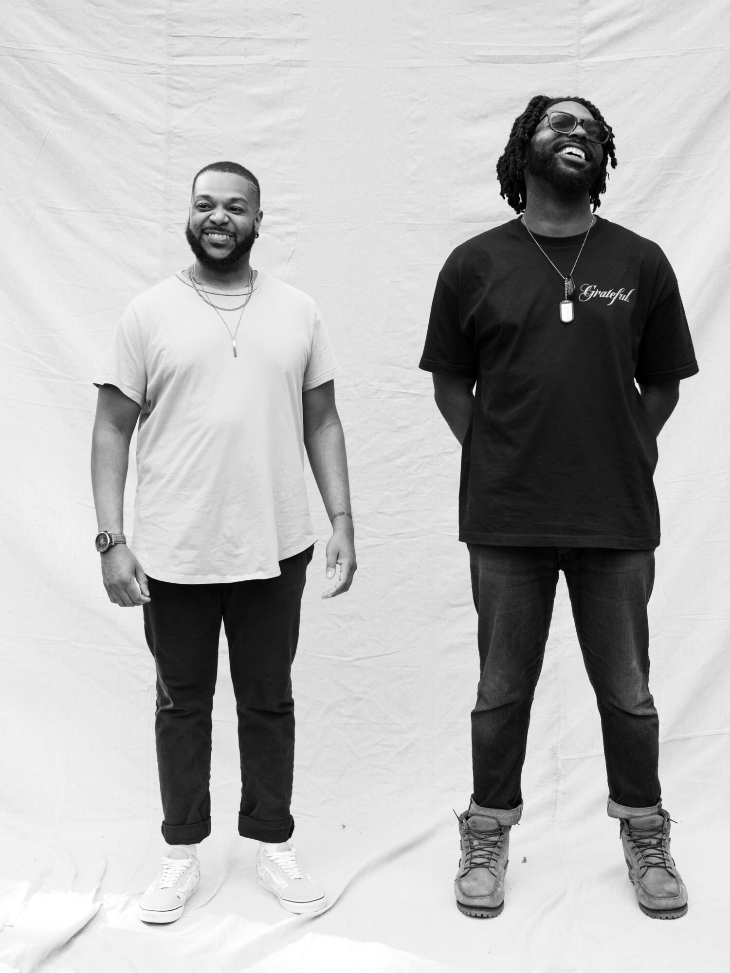 Two men standing side by side, smiling, against a plain background, one wearing a light t-shirt and the other a dark t-shirt.
