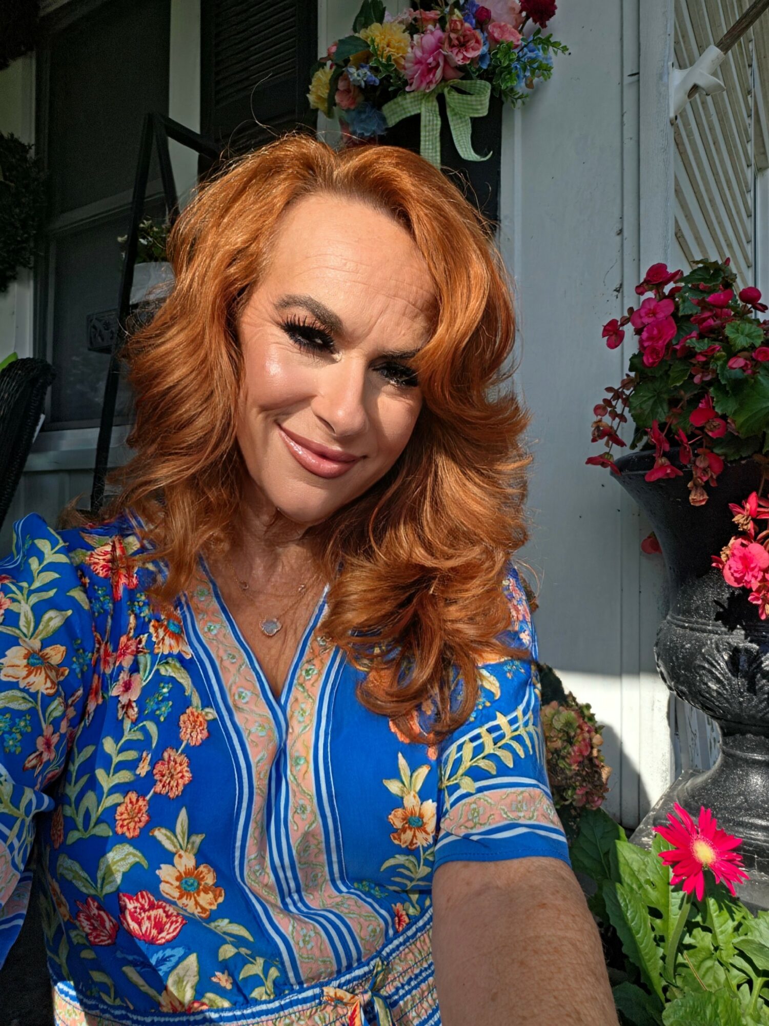 Woman with red hair smiling outdoors, surrounded by colorful flowers and plants.