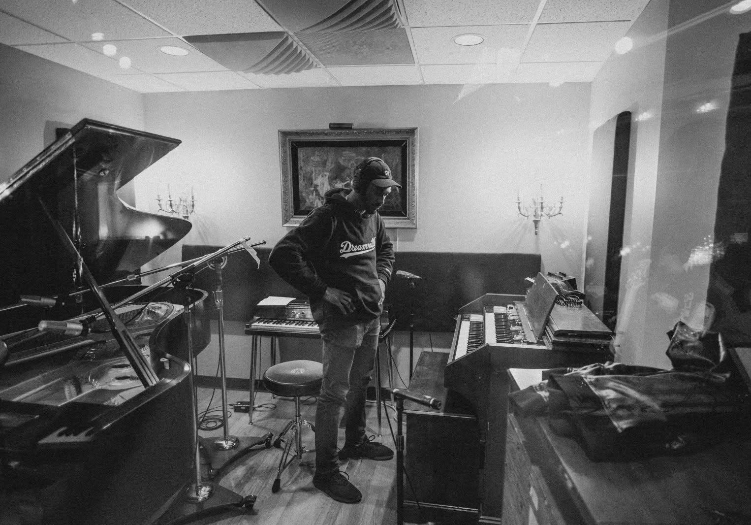 Person standing in a music studio with pianos, a painting on the wall, and a candelabra, in black and white.