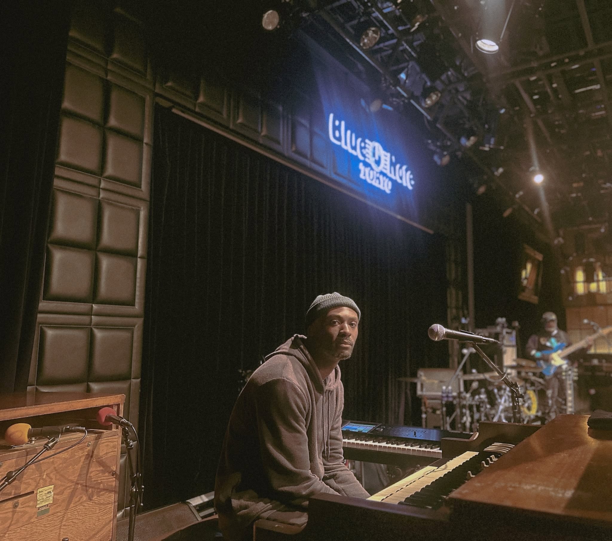 Person sitting at a keyboard on stage with a microphone, stage lights, and a band in the background.